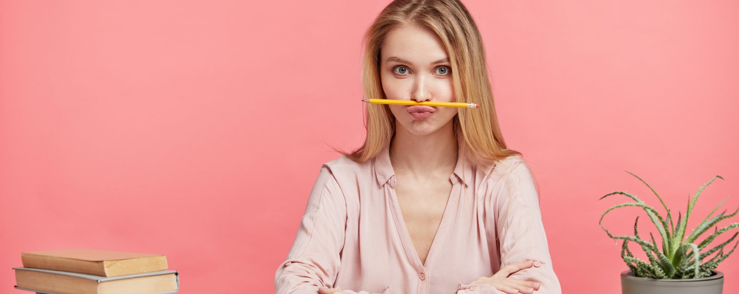 Horizontal portrait of funny female student keeps pencil on mouth, foolishes after long prepareation to pass course paper, surrounded with many books, isolated over pink background. Studying concept